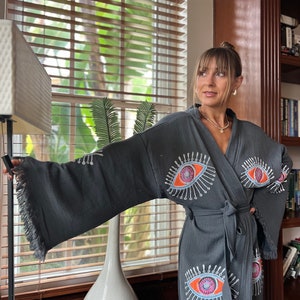 May include: A dark grey robe with a fringed hem and a tie waist. The robe features embroidered eye designs in orange, pink, and white. The sleeves are wide and the fabric appears to be textured.