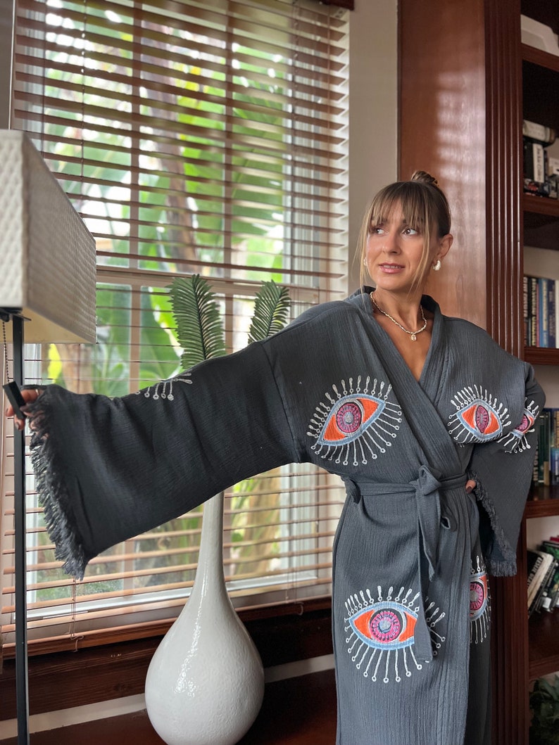 May include: A dark grey robe with a fringed hem and a tie waist. The robe features embroidered eye designs in orange, pink, and white. The sleeves are wide and the fabric appears to be textured.