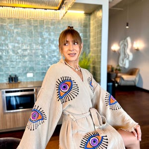 May include: A beige robe featuring embroidered eye motifs in orange, purple, and blue. The robe has a belt and fringing on the sleeves. The fabric of the robe is soft and textured.