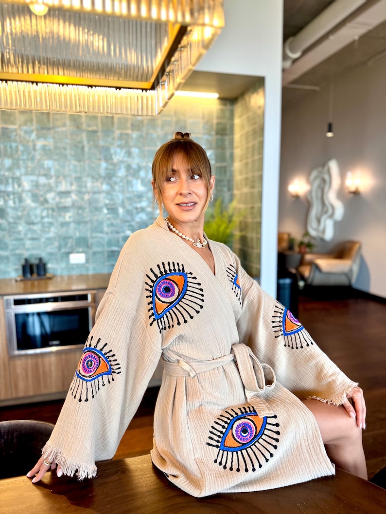 May include: A beige robe featuring embroidered eye motifs in orange, purple, and blue. The robe has a belt and fringing on the sleeves. The fabric of the robe is soft and textured.
