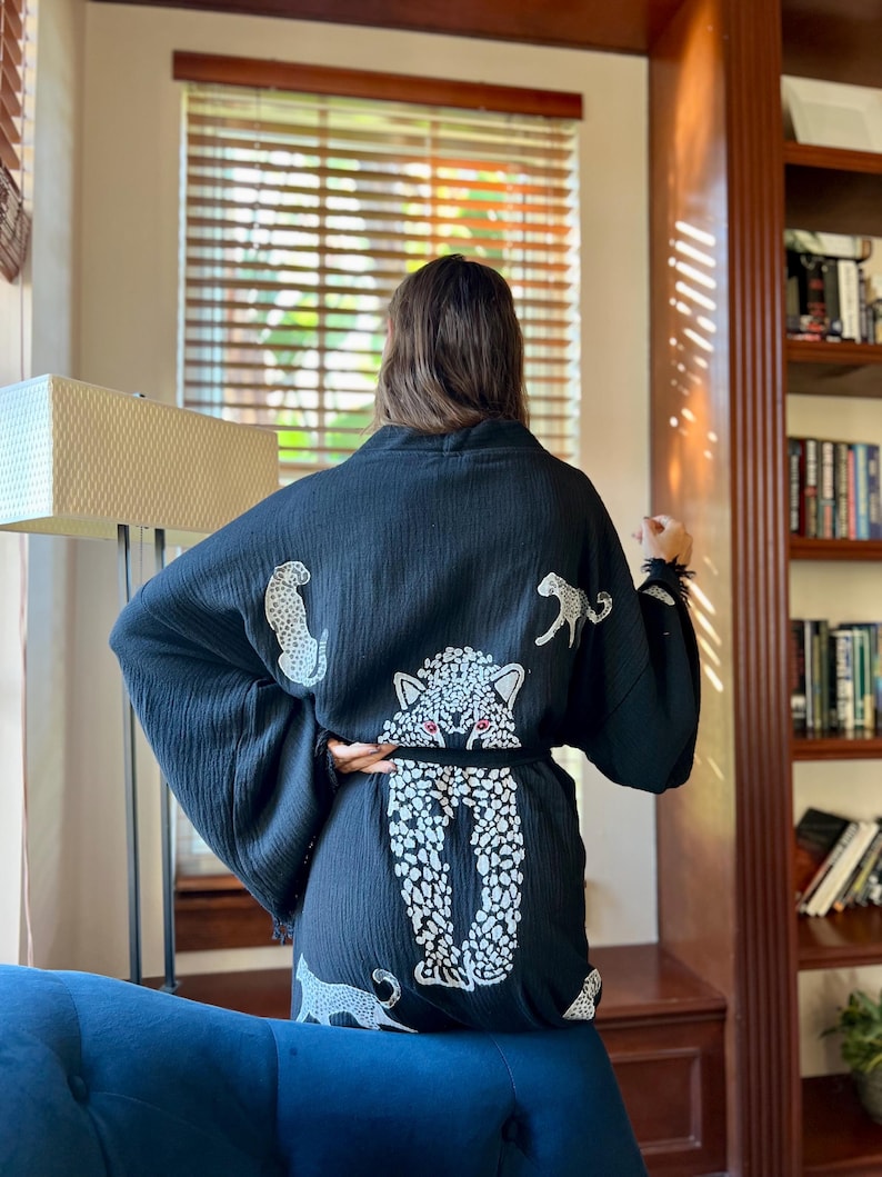 May include: Black kimono robe with white leopard print. The robe has a belt and a large leopard design on the back, with more leopard figures on the fabric. The sleeves are wide and flowing. The robe is made of a soft, textured material.