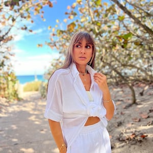 May include: A white linen outfit, consisting of a button-down shirt tied at the waist and matching pants. The outfit is accessorized with gold jewelry, including a necklace and bracelet. The background features a sandy path, trees, and a glimpse of the ocean.