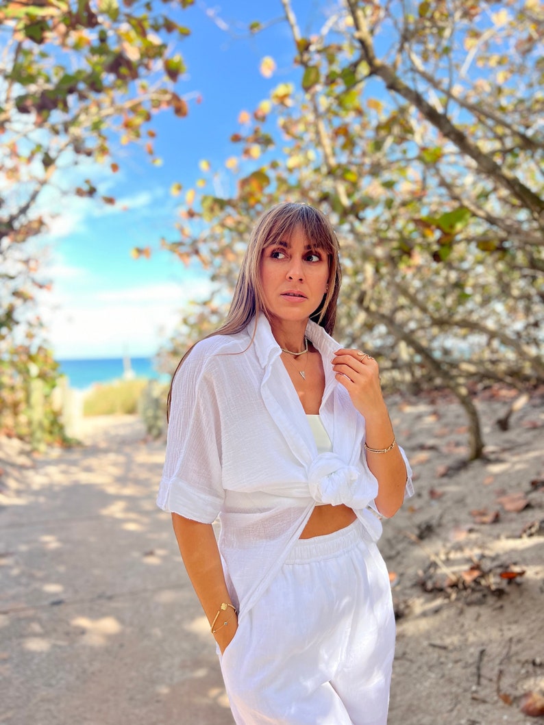 May include: A white linen outfit, consisting of a button-down shirt tied at the waist and matching pants. The outfit is accessorized with gold jewelry, including a necklace and bracelet. The background features a sandy path, trees, and a glimpse of the ocean.
