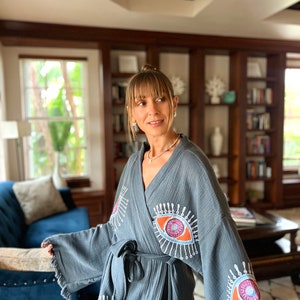 May include: A grey robe with an eye design. The robe has a tie waist and fringing on the sleeves. The eye design is in shades of orange, pink, and blue with white accents. The robe is made of a soft, textured fabric.