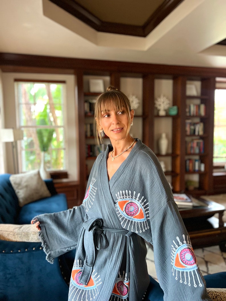 May include: A grey robe with an eye design. The robe has a tie waist and fringing on the sleeves. The eye design is in shades of orange, pink, and blue with white accents. The robe is made of a soft, textured fabric.