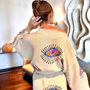 May include: Beige, off-the-shoulder kimono-style top and matching trousers set. The set features embroidered eye designs in blue, orange, and purple. The kimono has fringed edges. The outfit is made of a lightweight, textured fabric.