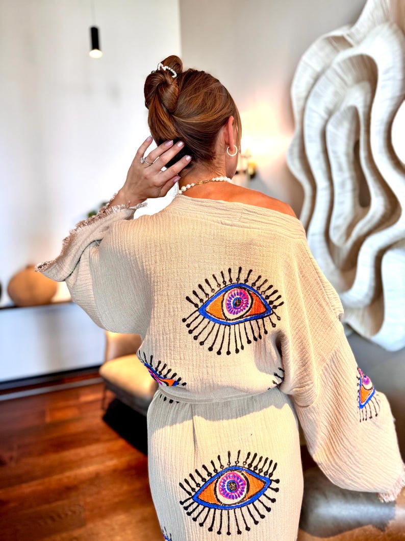May include: Beige, off-the-shoulder kimono-style top and matching trousers set. The set features embroidered eye designs in blue, orange, and purple. The kimono has fringed edges. The outfit is made of a lightweight, textured fabric.