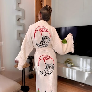 K&ouml;nnte beinhalten: Eine wei&szlig;e Baumwollrobe mit Fransenbesatz zeigt gestickte Weihnachtsmotive. Die Robe hat auf der R&uuml;ckseite ein rotes und schwarzes Weihnachtsmann-Motiv und unten ein gr&uuml;nes Weihnachtsbaum- und Rentier-Motiv.
