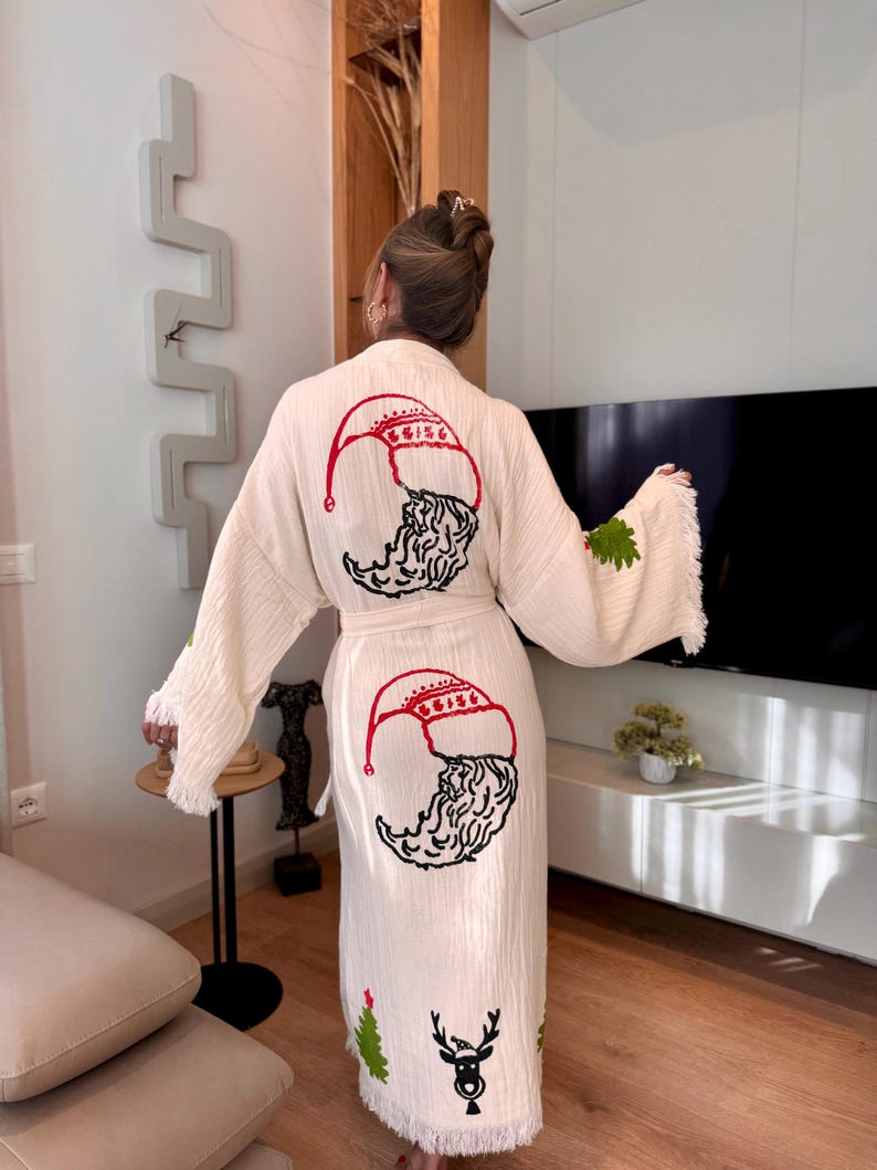 K&ouml;nnte beinhalten: Eine wei&szlig;e Baumwollrobe mit Fransenbesatz zeigt gestickte Weihnachtsmotive. Die Robe hat auf der R&uuml;ckseite ein rotes und schwarzes Weihnachtsmann-Motiv und unten ein gr&uuml;nes Weihnachtsbaum- und Rentier-Motiv.