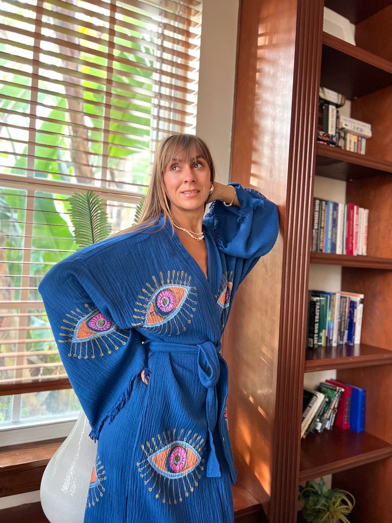 May include: A blue robe featuring an eye pattern. The robe has a tie waist and fringed sleeves. The eye design is embroidered with pink, orange, and gold thread. The robe is made of a textured fabric.