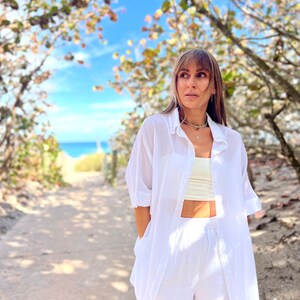 May include: A woman wearing a white linen shirt, a cream-colored top, and white linen pants. The outfit is paired with a delicate silver necklace. The background features a pathway leading to the beach, framed by trees and a clear blue sky.