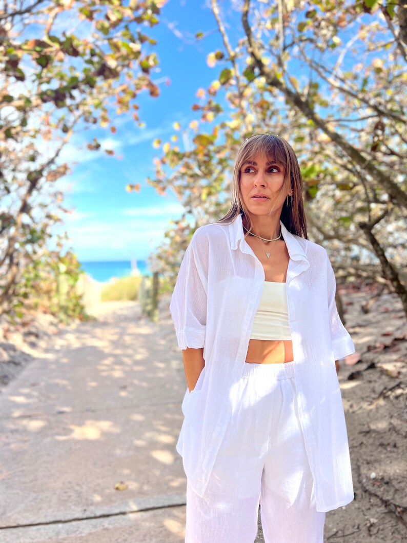 May include: A woman wearing a white linen shirt, a cream-colored top, and white linen pants. The outfit is paired with a delicate silver necklace. The background features a pathway leading to the beach, framed by trees and a clear blue sky.