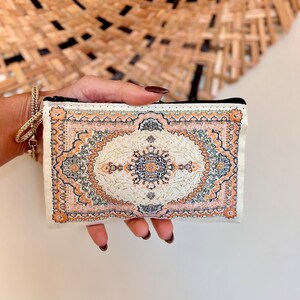 May include: A small white zippered pouch with a colorful patterned design resembling a rug. The pattern features shades of orange, green, and brown.