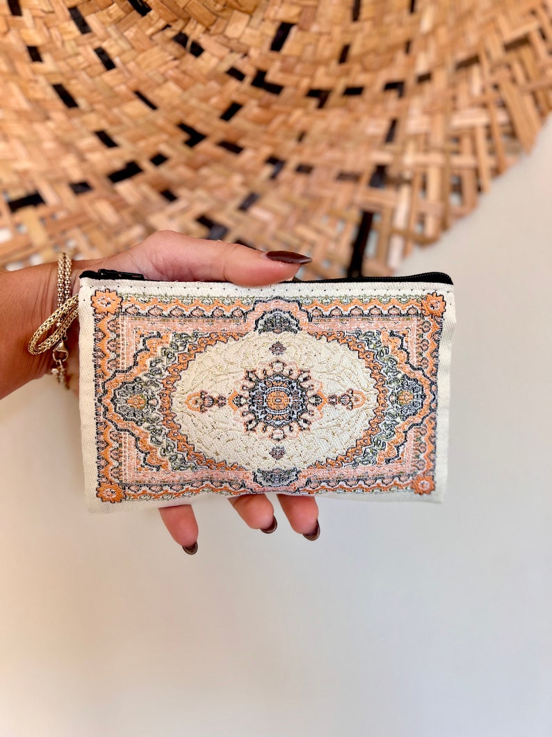 May include: A small white zippered pouch with a colorful patterned design resembling a rug. The pattern features shades of orange, green, and brown.