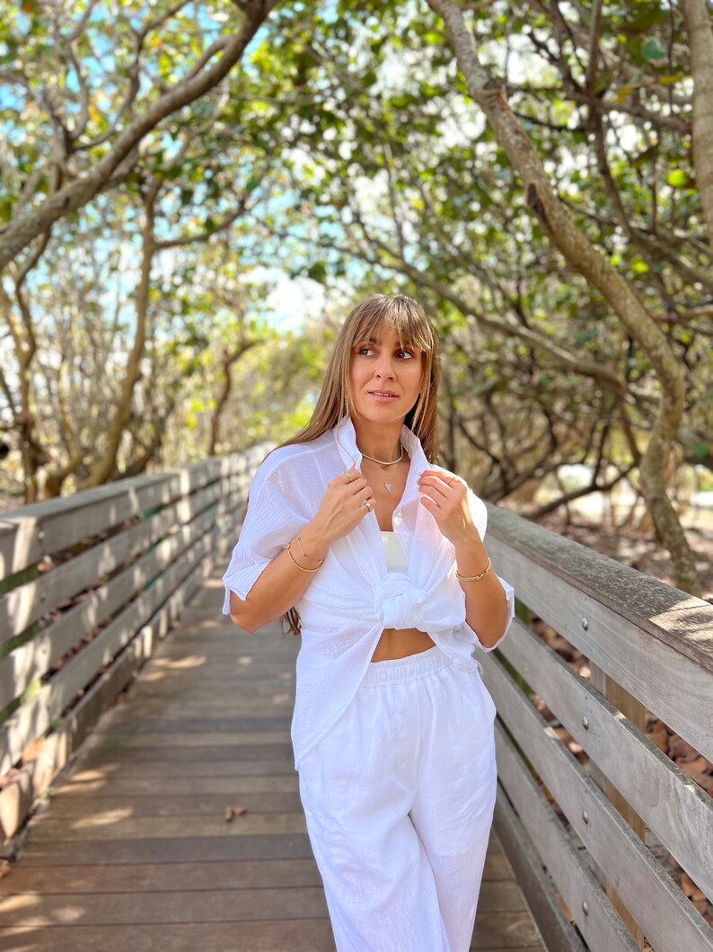 May include: A white linen outfit, consisting of a button-down shirt tied at the waist and matching wide-leg pants. The outfit is worn on a wooden boardwalk with trees in the background. The shirt has short sleeves.