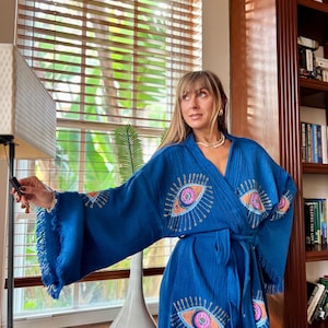 May include: A blue robe featuring embroidered eye motifs on the sleeves and front. The robe has a fringed hem and a matching belt. The eye designs are in pink, orange, and blue, with gold detailing. The fabric has a textured appearance.