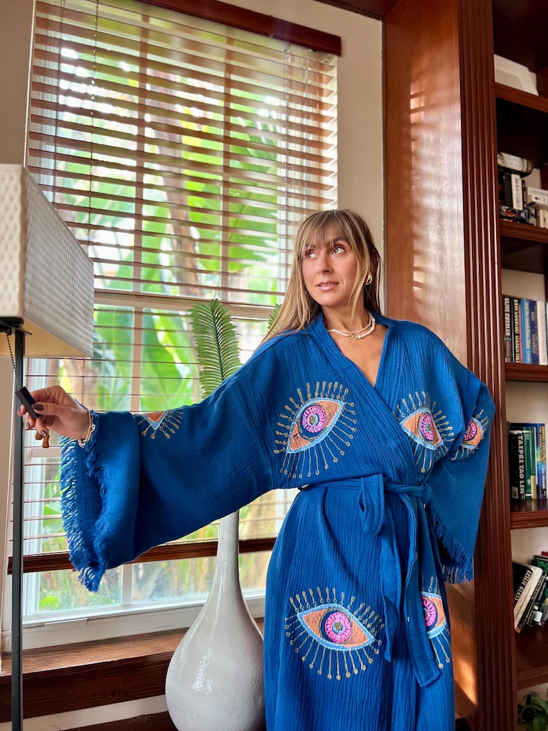 May include: A blue robe featuring embroidered eye motifs on the sleeves and front. The robe has a fringed hem and a matching belt. The eye designs are in pink, orange, and blue, with gold detailing. The fabric has a textured appearance.