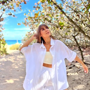 May include: A white linen shirt and matching pants set. The shirt is button-down with short sleeves, and the pants have an elastic waistband. The outfit is paired with a cream-colored crop top and gold jewelry. The background features a beach and blue sky.