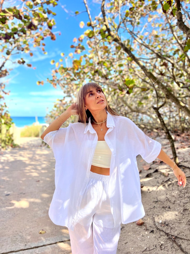 May include: A white linen shirt and matching pants set. The shirt is button-down with short sleeves, and the pants have an elastic waistband. The outfit is paired with a cream-colored crop top and gold jewelry. The background features a beach and blue sky.