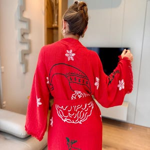 K&ouml;nnte beinhalten: Rote Weihnachtsrobe mit wei&szlig;en gestickten Schneeflocken, einem Rentier und einem Weihnachtsmann-Gesicht. Die Robe hat eine lange, flie&szlig;ende Silhouette und einen Bindeg&uuml;rtel.