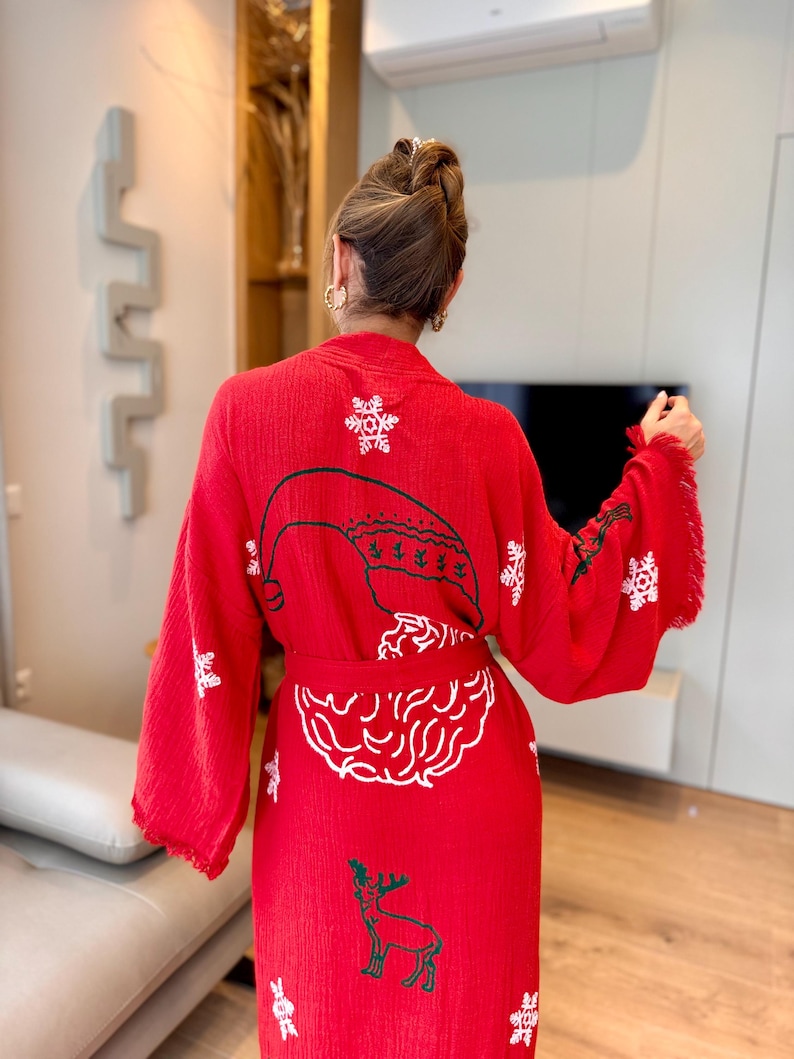 K&ouml;nnte beinhalten: Rote Weihnachtsrobe mit wei&szlig;en gestickten Schneeflocken, einem Rentier und einem Weihnachtsmann-Gesicht. Die Robe hat eine lange, flie&szlig;ende Silhouette und einen Bindeg&uuml;rtel.