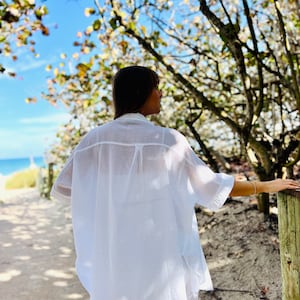 May include: A white, oversized button-down shirt with short sleeves. The shirt is made of a lightweight, textured fabric. The person is standing on a sandy path near the beach, with trees and a blue sky in the background.
