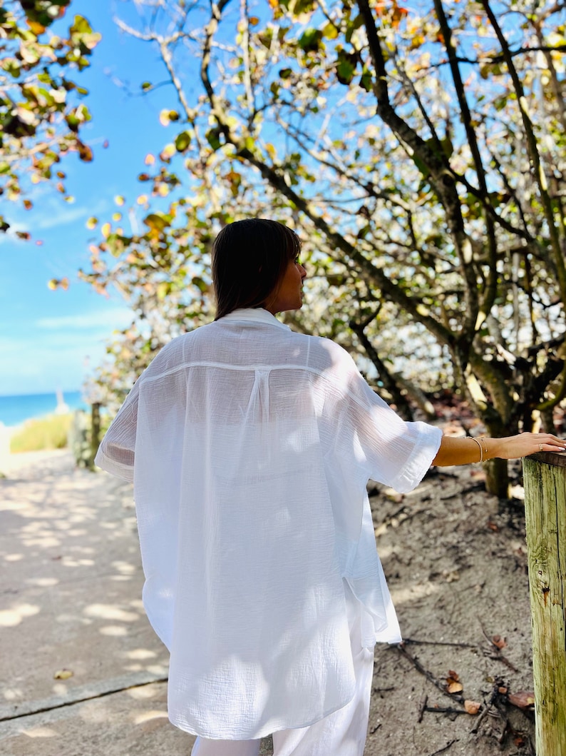 May include: A white, oversized button-down shirt with short sleeves. The shirt is made of a lightweight, textured fabric. The person is standing on a sandy path near the beach, with trees and a blue sky in the background.
