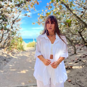 May include: A white linen shirt and matching pants set. The shirt is open, revealing a white crop top. The outfit is accessorized with gold jewelry. The background features a pathway leading to the ocean under a blue sky.
