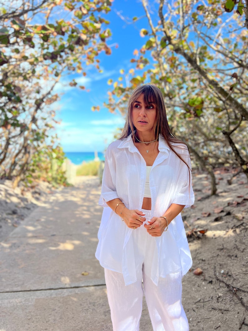 May include: A white linen shirt and matching pants set. The shirt is open, revealing a white crop top. The outfit is accessorized with gold jewelry. The background features a pathway leading to the ocean under a blue sky.