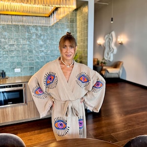 May include: Beige robe featuring embroidered eye designs in orange, purple, and blue. The robe has a fringed hem and a matching tie belt. The background includes a kitchen and a living room.