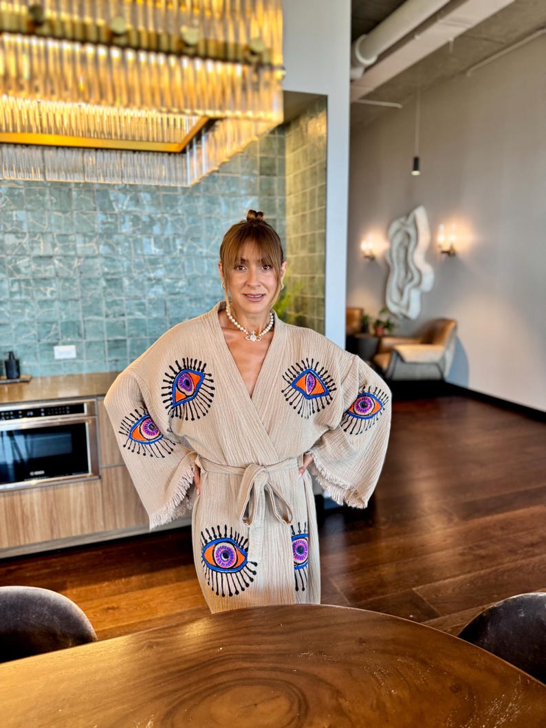 May include: Beige robe featuring embroidered eye designs in orange, purple, and blue. The robe has a fringed hem and a matching tie belt. The background includes a kitchen and a living room.