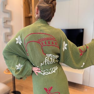 K&ouml;nnte beinhalten: Ein gr&uuml;nes Gewand mit wei&szlig;en und roten gestickten Schneeflocken, einem Hirsch und einem Weihnachtsbaumdesign. Das Gewand hat einen Bindeg&uuml;rtel und Fransenbesatz.