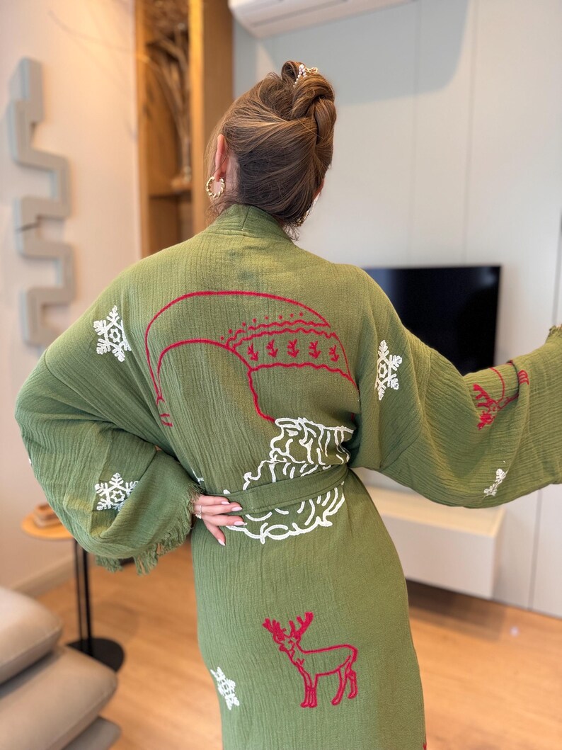 K&ouml;nnte beinhalten: Ein gr&uuml;nes Gewand mit wei&szlig;en und roten gestickten Schneeflocken, einem Hirsch und einem Weihnachtsbaumdesign. Das Gewand hat einen Bindeg&uuml;rtel und Fransenbesatz.