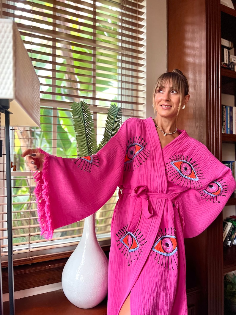 May include: A bright pink robe with an eye pattern in orange, blue, and black. The wrap-around robe has a matching belt and fringed sleeves. The fabric has a textured appearance. The robe is a stylish garment.