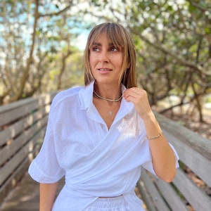 May include: A woman wearing a white wrap shirt and matching pants, accessorized with gold jewelry. The outfit is styled with a delicate necklace and bracelets. The background features a wooden bridge and lush greenery, creating a natural setting.