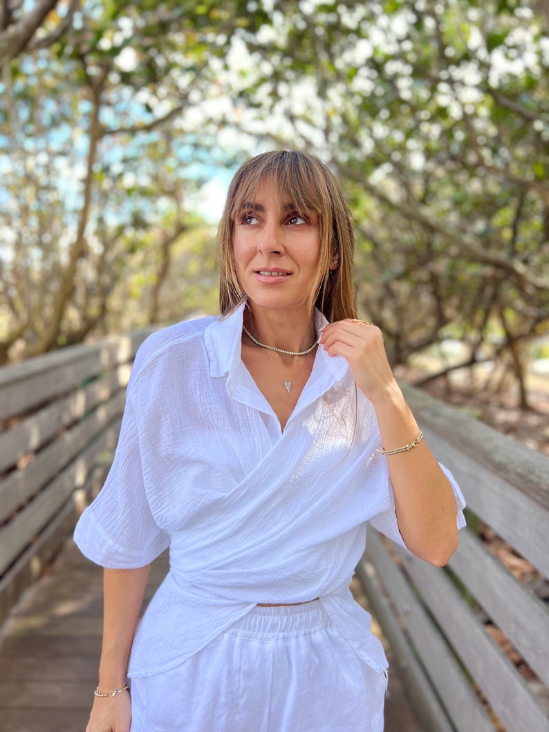 May include: A woman wearing a white wrap shirt and matching pants, accessorized with gold jewelry. The outfit is styled with a delicate necklace and bracelets. The background features a wooden bridge and lush greenery, creating a natural setting.