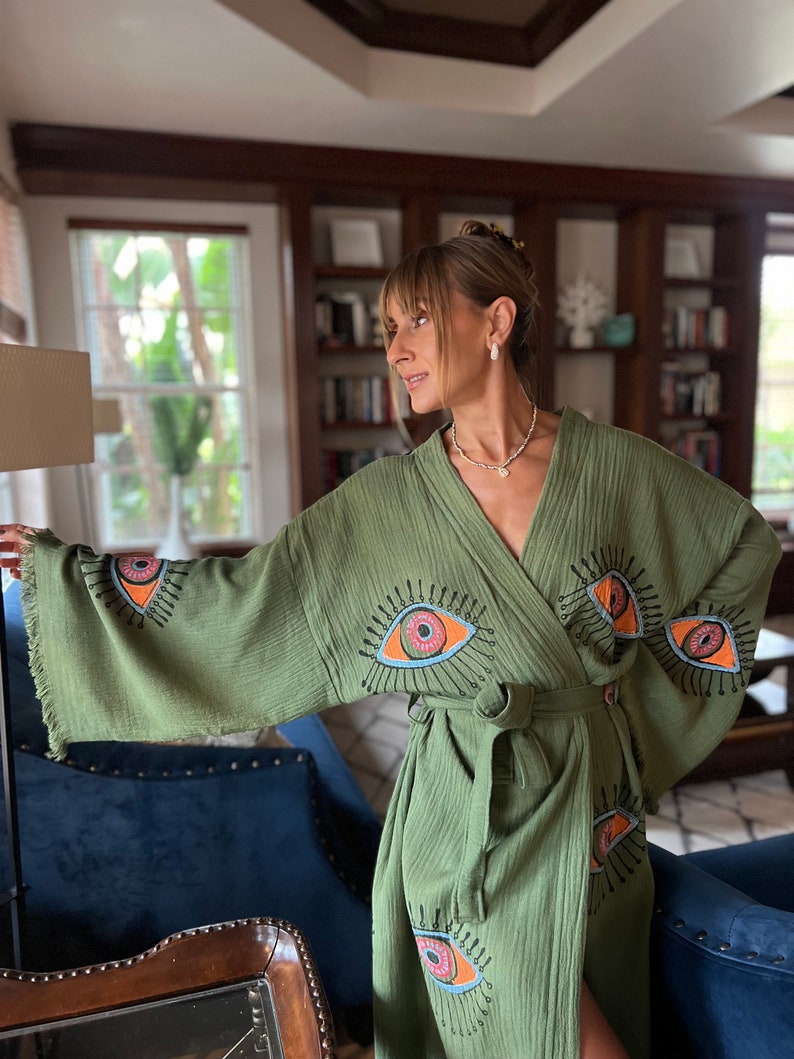 May include: An olive green robe with an eye design. The robe has a wrap-around style and a matching belt. The eye design is in orange, pink, and blue. The sleeves have a fringed edge.