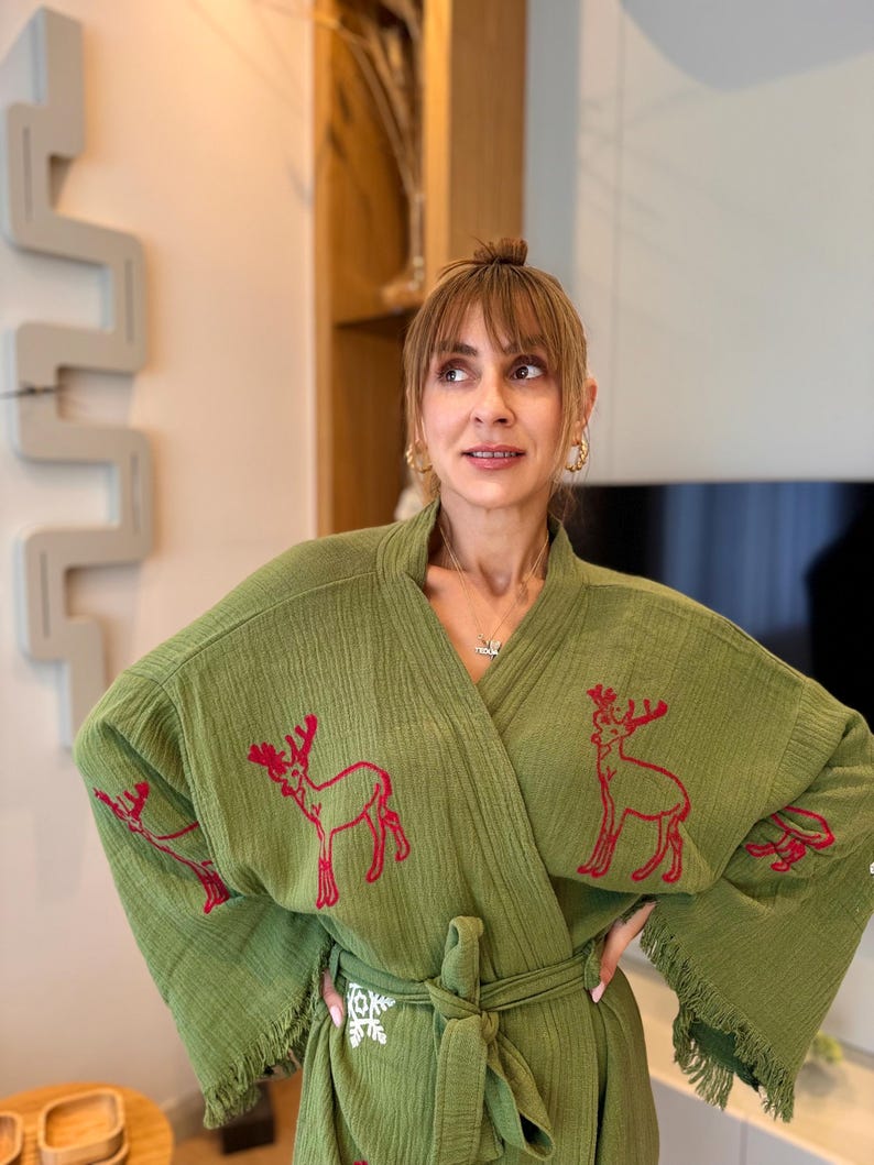 K&ouml;nnte beinhalten: Eine gr&uuml;ne Baumwollrobe mit roten gestickten Rentieren und einem wei&szlig;en Schneeflockenmuster. Die Robe hat einen Bindeg&uuml;rtel und Fransenbesatz.