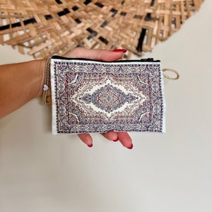 May include: A small white zippered pouch with a colourful rug pattern. The pouch is held in a hand with red nail polish.