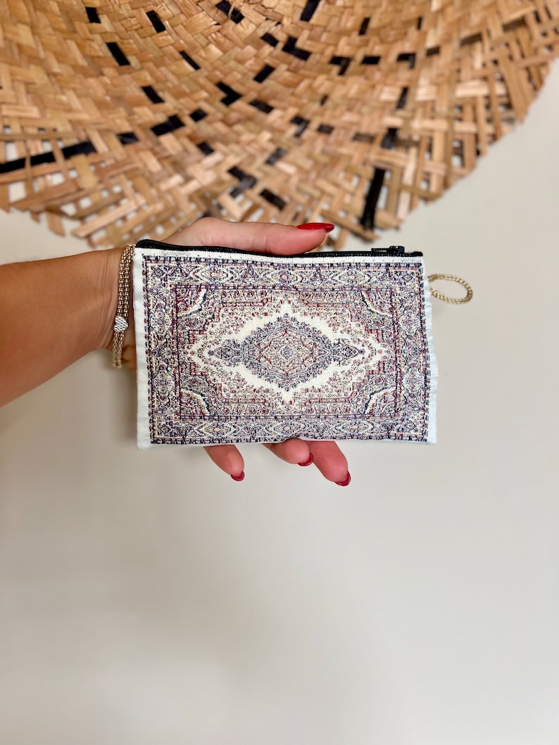 May include: A small white zippered pouch with a colourful rug pattern. The pouch is held in a hand with red nail polish.