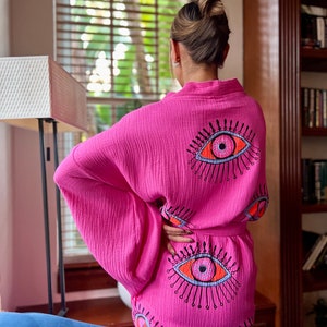 May include: A vibrant pink kimono-style robe with an eye motif. The robe has a wrap-around design and a matching belt. The eye design is embroidered in orange, black, and blue. The robe is made of a lightweight, textured fabric.
