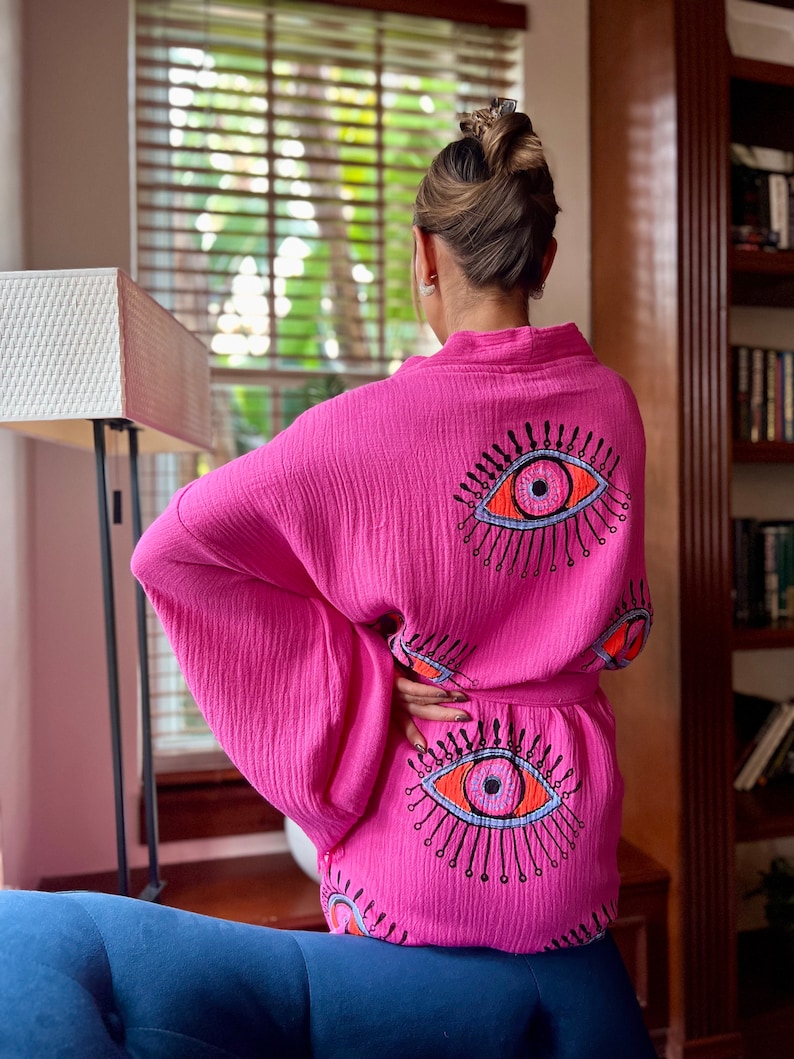 May include: A vibrant pink kimono-style robe with an eye motif. The robe has a wrap-around design and a matching belt. The eye design is embroidered in orange, black, and blue. The robe is made of a lightweight, textured fabric.