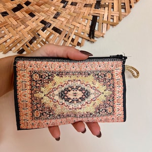 May include: A small black zippered pouch with a colorful rug design. The rug design features a central diamond shape with gold accents.