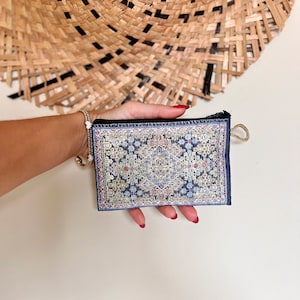 May include: A small blue zippered pouch with a colorful patterned design reminiscent of a rug. The pouch is held in a hand with red nail polish.