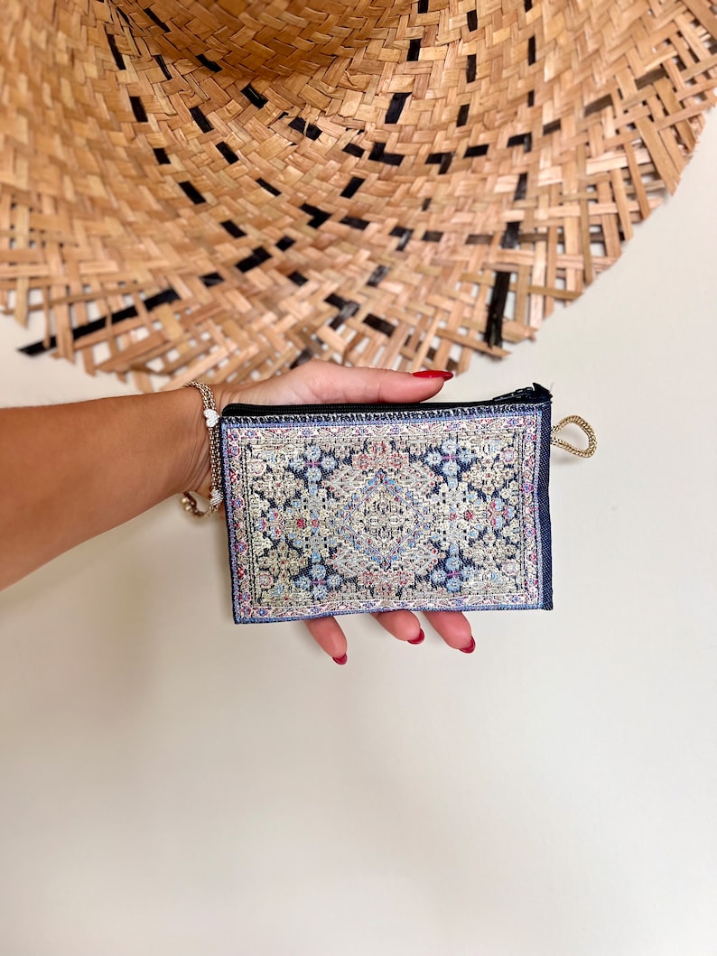 May include: A small blue zippered pouch with a colorful patterned design reminiscent of a rug. The pouch is held in a hand with red nail polish.