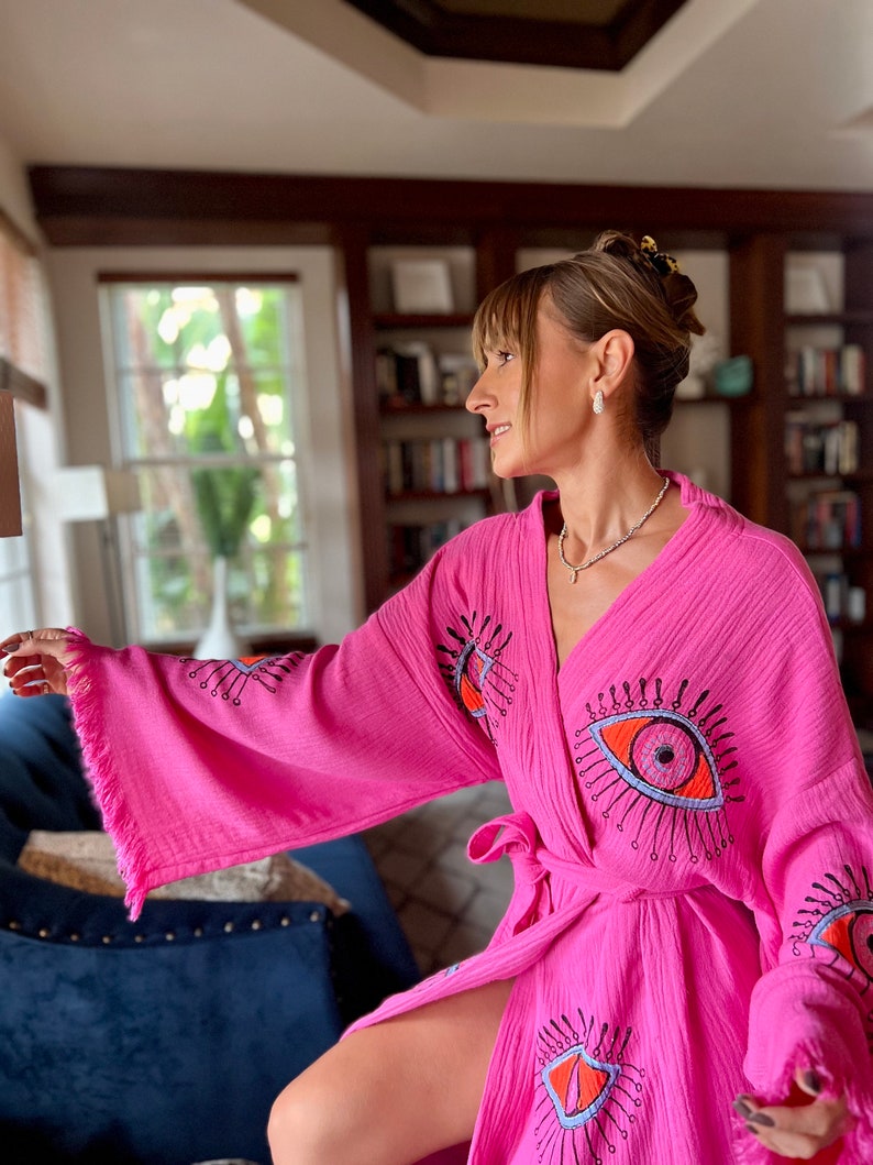 May include: A vibrant pink robe with a wrap-around style. The robe is adorned with large eye motifs in orange, blue, and black. The sleeves have a fringed trim. It is secured at the waist with a matching belt.