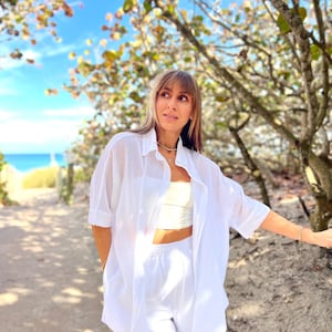 May include: A white linen shirt and matching shorts set. The shirt is buttoned and has short sleeves. The shorts are high-waisted. The outfit is worn with a cream-colored crop top and gold jewelry. The background includes a beach and blue sky.