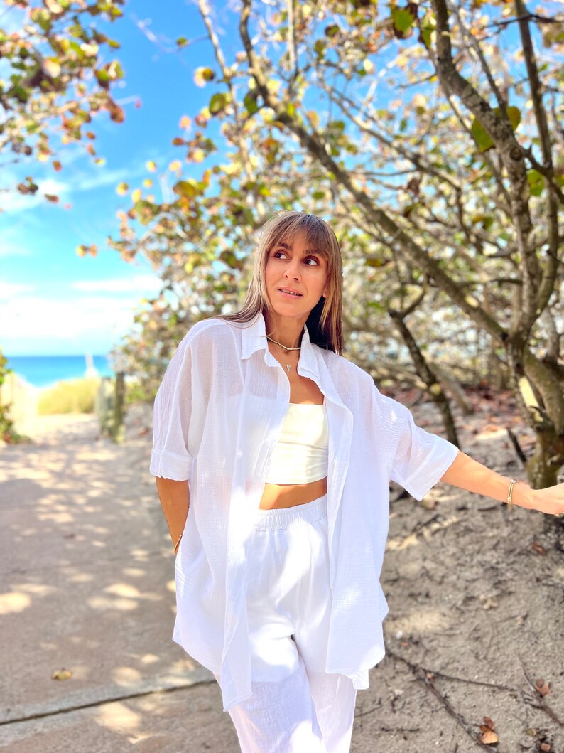 May include: A white linen shirt and matching shorts set. The shirt is buttoned and has short sleeves. The shorts are high-waisted. The outfit is worn with a cream-colored crop top and gold jewelry. The background includes a beach and blue sky.