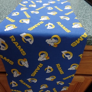 May include: A blue table runner featuring a repeating pattern of the word "RAMS" and the team's logo in white and yellow. The runner is made of fabric and is displayed on a countertop.