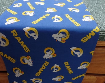 May include: A blue table runner featuring a repeating pattern of the word "RAMS" and the team's logo in white and yellow. The runner is made of fabric and is displayed on a countertop.